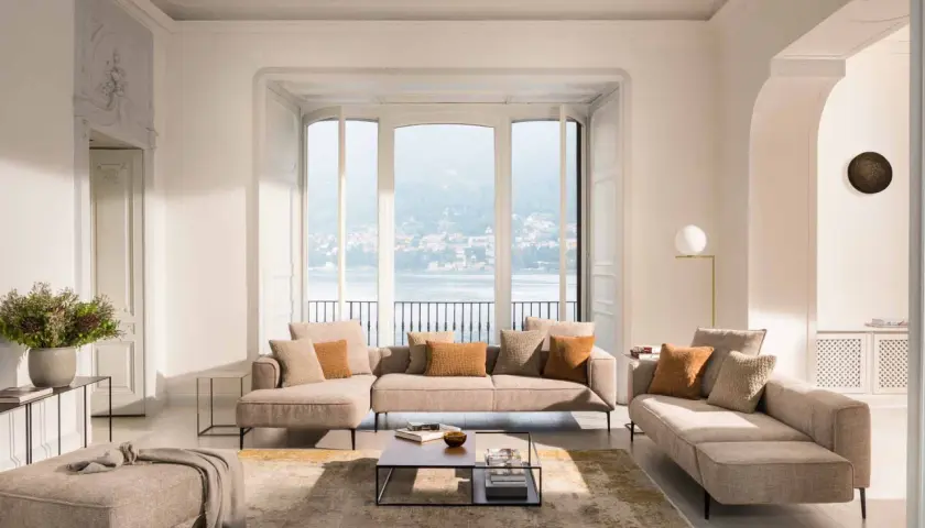 Un salon cocooning moderne avec canapé modulable, pouf, tapis et vue sur un lac.