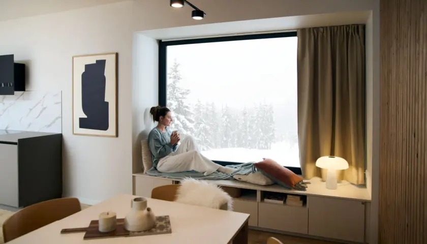 Un coin lecture scandinave parfait pour admirer la neige. Une femme savoure une boisson chaude, enveloppée dans un plaid douillet.