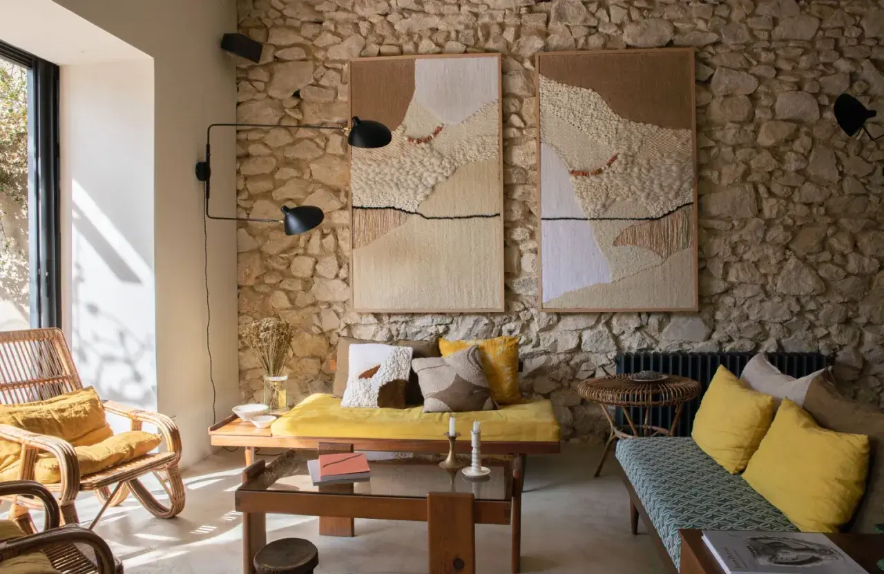Un salon ancien moderne avec des tapisseries texturées sur un mur de pierre, des meubles en rotin et des coussins jaune vif.