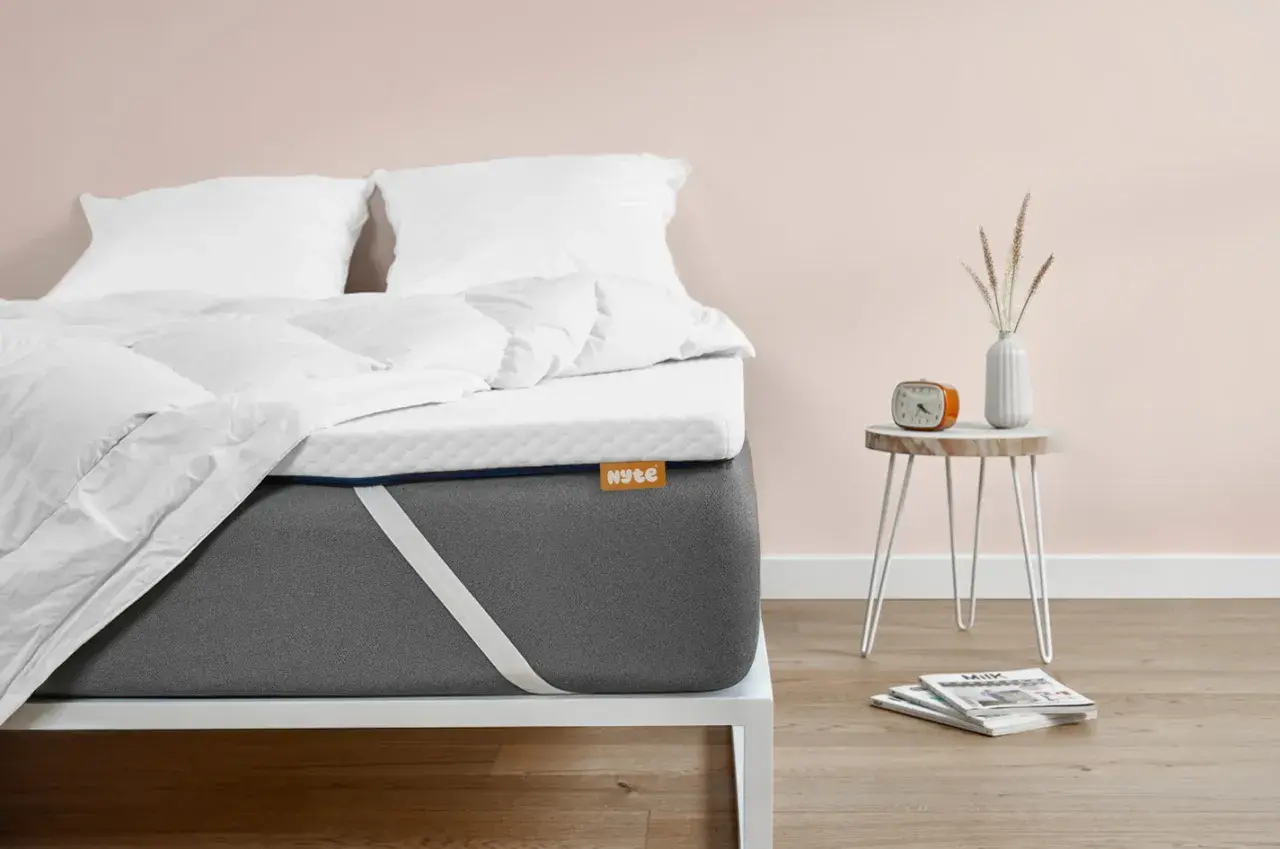Un matelas trop mou que faire ? Ce surmatelas blanc sur un matelas gris offre une solution. Un réveil orange et des magazines sont sur une table de chevet.