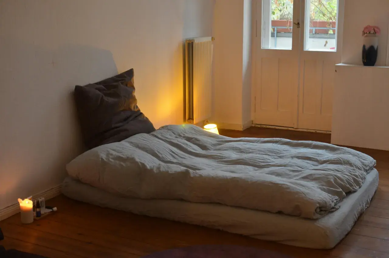 Un matelas au sol sans sommier, prêt pour une nuit paisible. Une bougie allumée éclaire doucement la pièce.