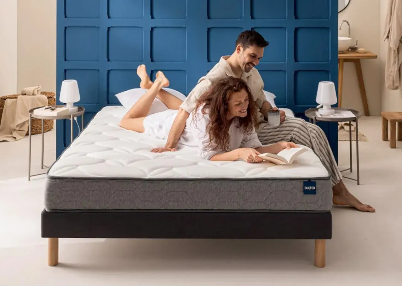 Un couple profite d'un moment de détente sur un matelas de grande taille. La femme lit un livre, l'homme boit une tasse.