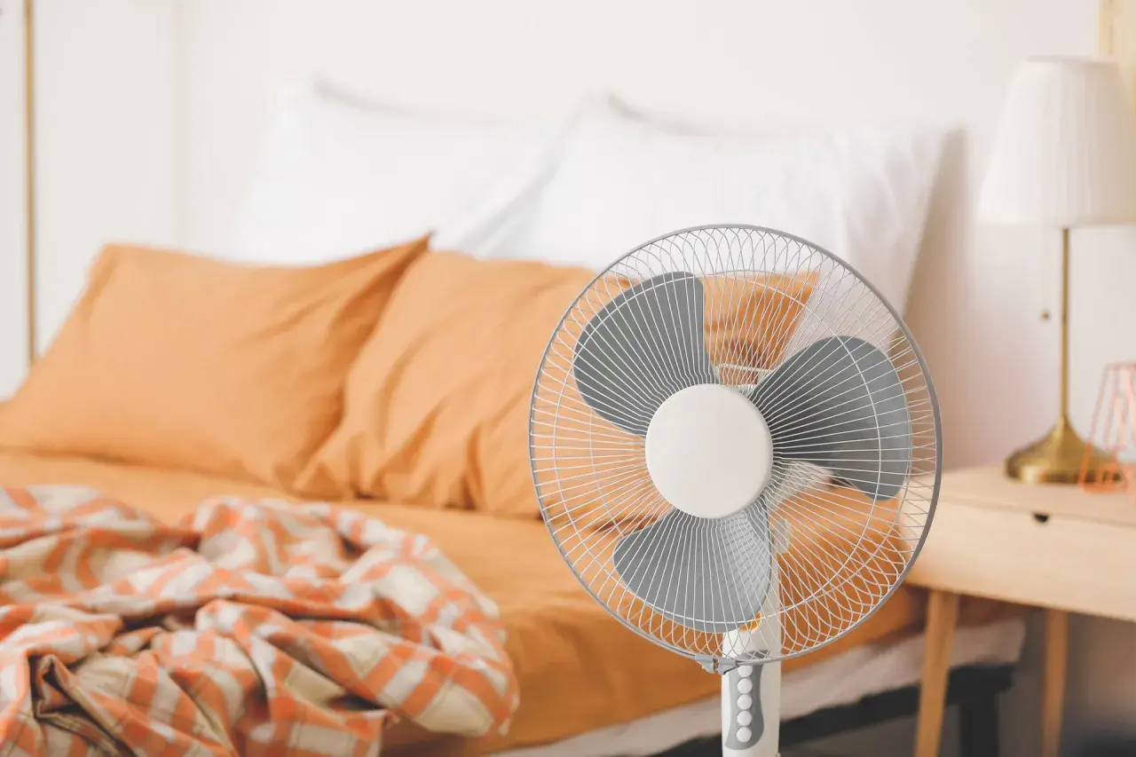Ventilateur sur pied prêt à rafraîchir une pièce, avec un lit défait en arrière-plan.