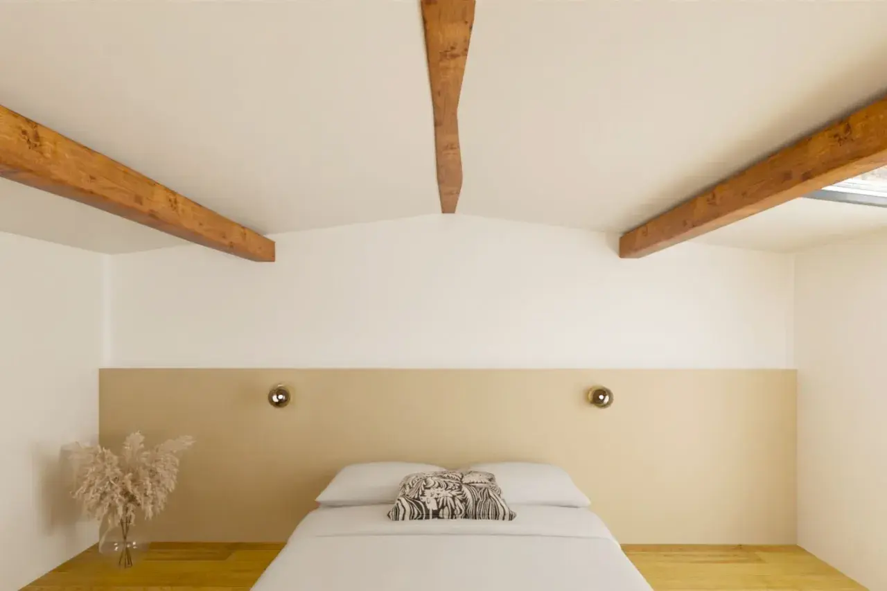 Chambre mansardée aux poutres apparentes, peinte en deux couleurs : blanc et beige. Le lit est fait, avec un coussin décoratif.