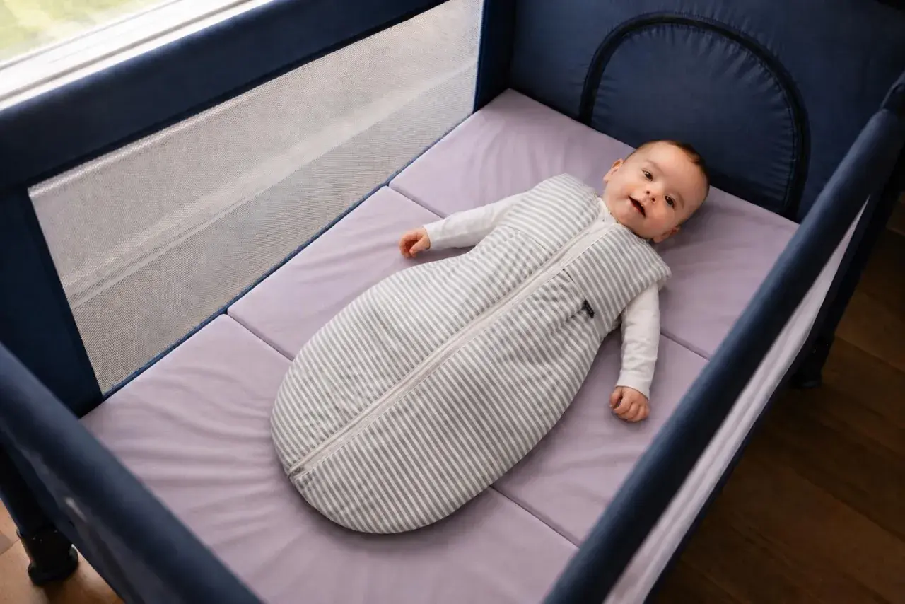 Bébé souriant dans un lit parapluie, sur un matelas adapté. La taille du matelas lit parapluie est parfaite pour le confort de bébé.