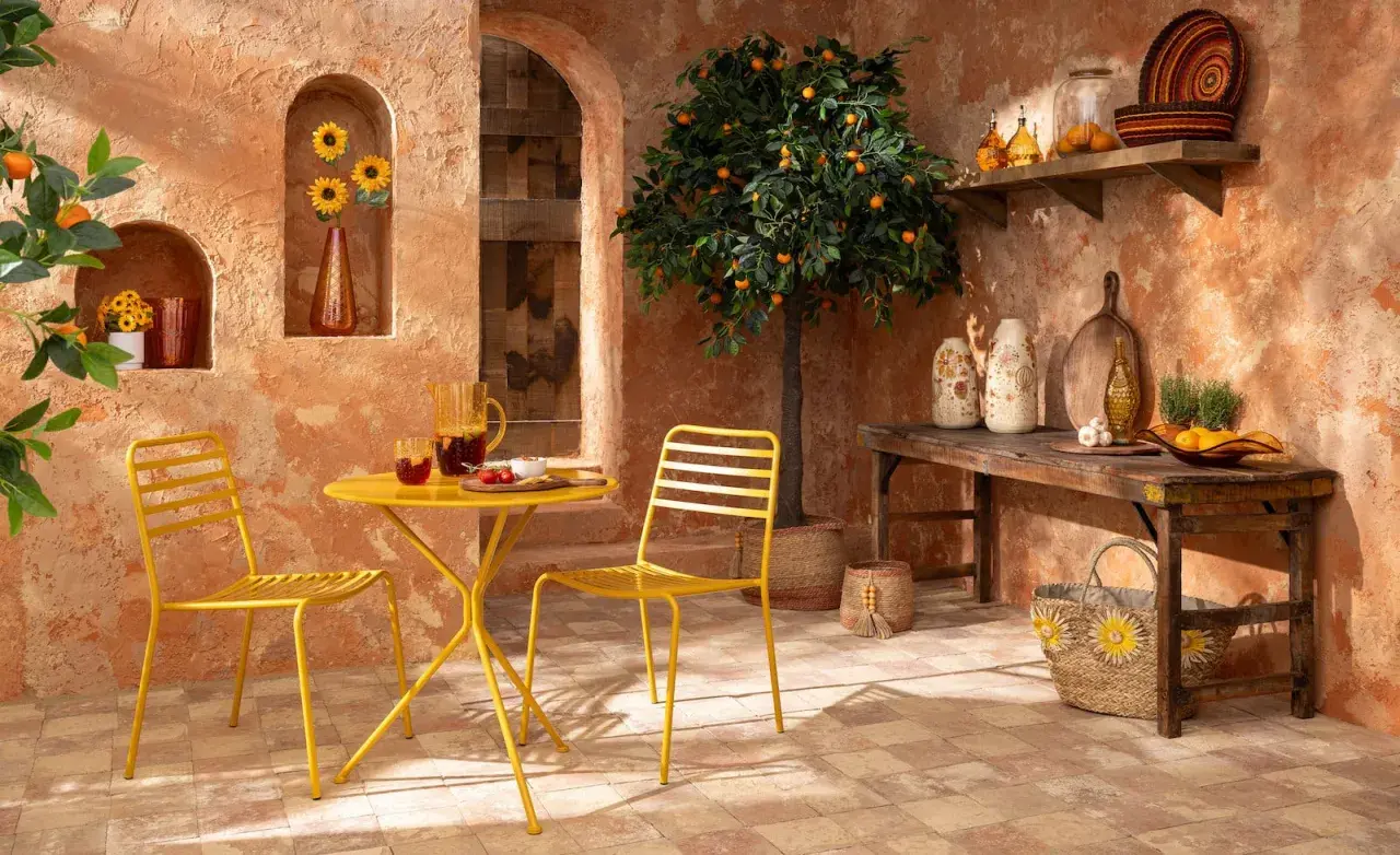 Terrasse ensoleillée aux couleurs terracotta et jaune. Table et chaises jaunes, oranger, tournesols et vaisselle en verre.