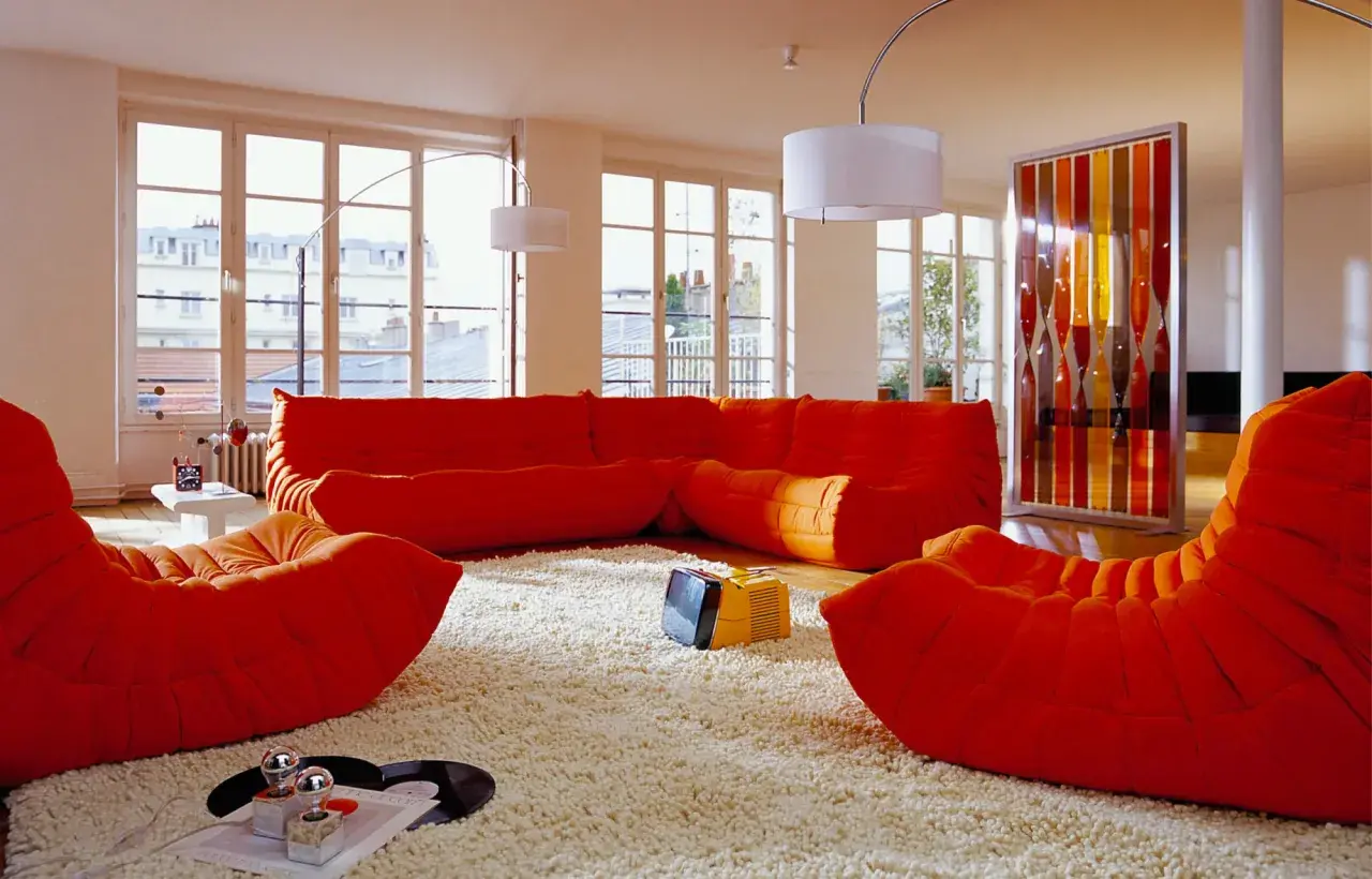 Salon aux couleurs vives, mobilier années 70 caractéristiques : canapé orange, fauteuils Togo, tapis shaggy, écran décoratif coloré.