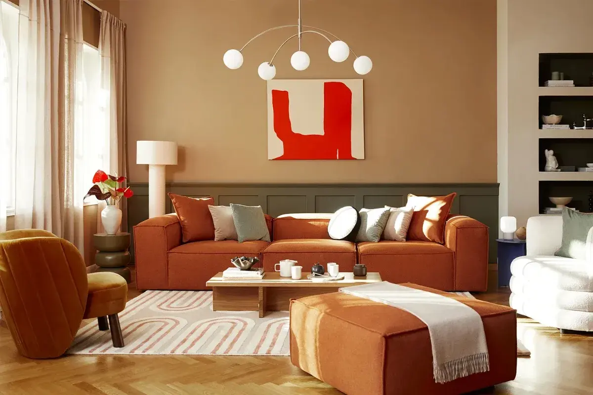 Salon cosy aux tons terreux. Un canapé orange vif, des coussins doux et une peinture abstraite rouge créent une ambiance chaleureuse. La **couleur chaude peinture** domine, invitant à la détente.