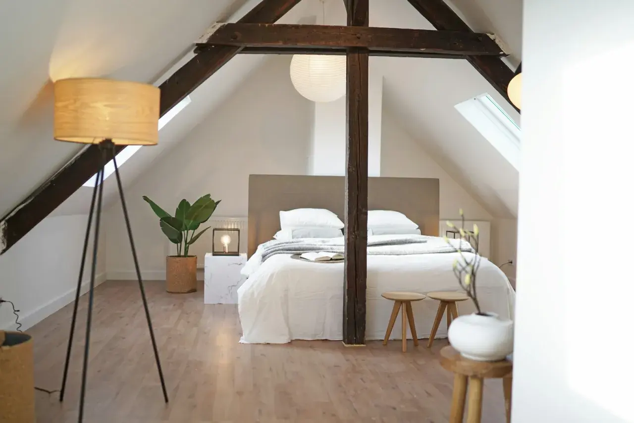 Une chambre minimaliste sous les combles, avec un lit blanc douillet, des poutres apparentes et une plante verte.