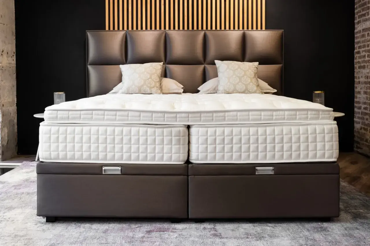 Un lit moderne avec un sommier capitonné marron, idéal pour poser un sommier à lattes. Le matelas est blanc et deux coussins décoratifs sont présents.