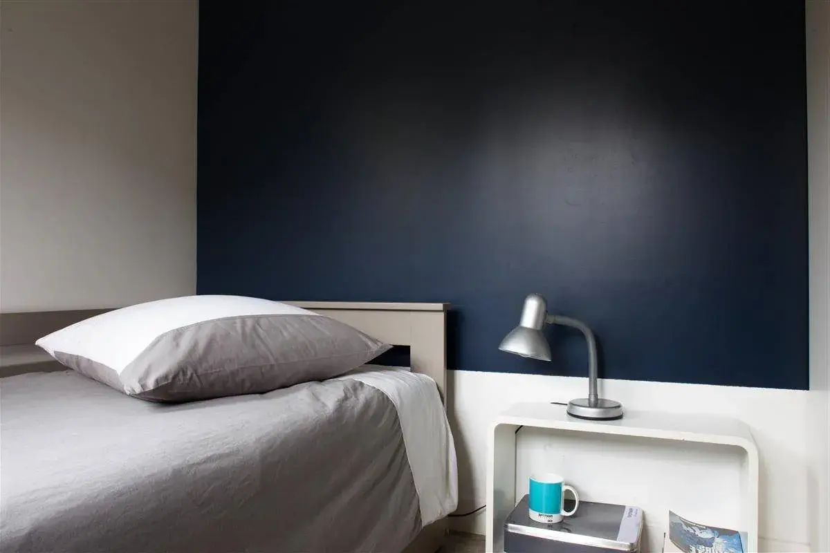 Chambre bleu nuit et taupe : un lit douillet avec un oreiller blanc et gris, une lampe de bureau argentée et une tasse bleue sur une table de chevet blanche.