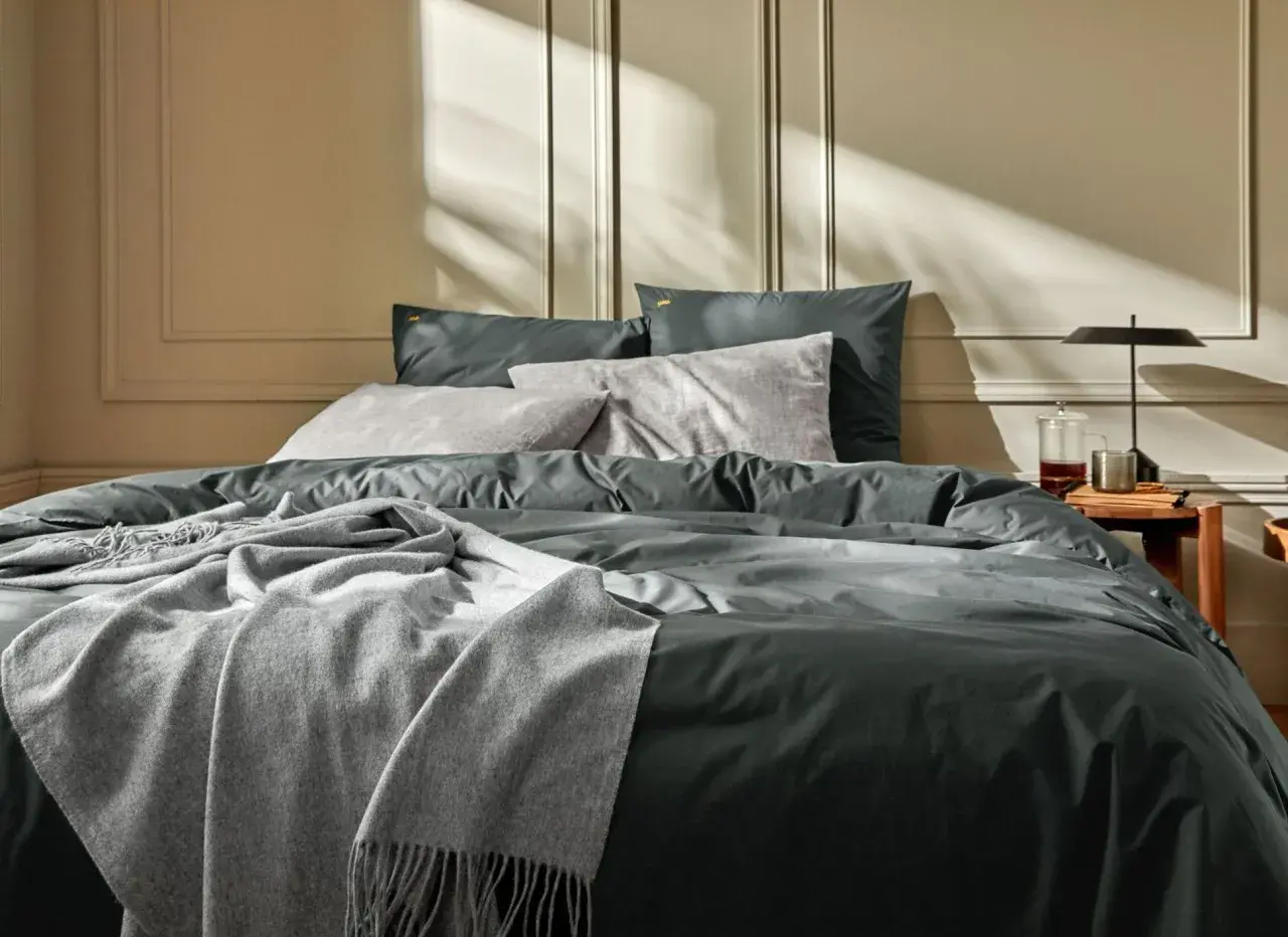 Un lit douillet avec un drap en soie ou satin anthracite, des coussins gris et une couverture douce. La lumière du soleil crée des ombres sur le mur.