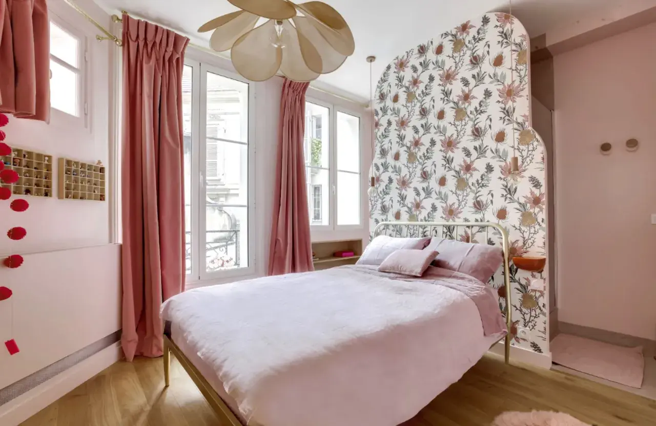 Chambre rose poudré et bois, avec un lit douillet, des rideaux velours, un papier peint floral et un lustre original.