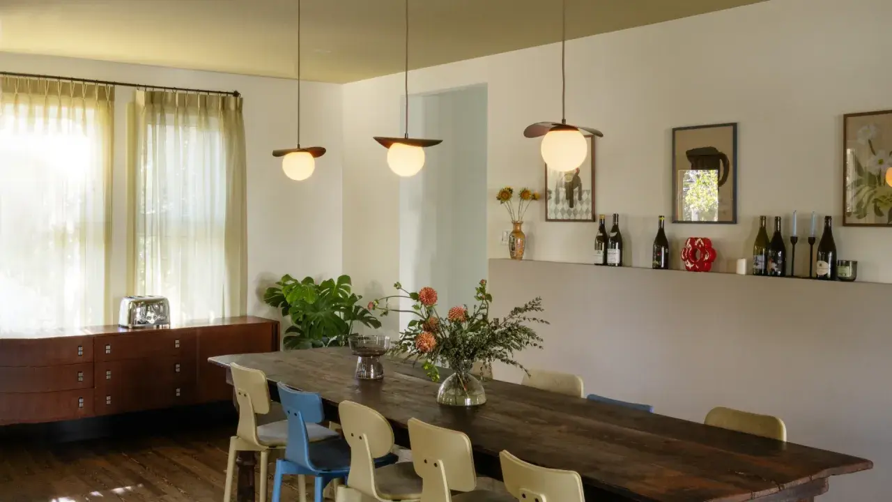 Trois luminaires au-dessus d'une grande table en bois, éclairant un bouquet de fleurs et des chaises colorées.