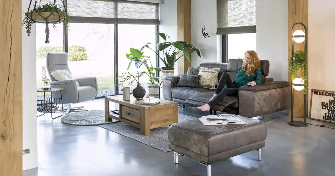 Une femme se détend dans un salon cocooning, avec un fauteuil confortable, un canapé en cuir et beaucoup de plantes vertes.