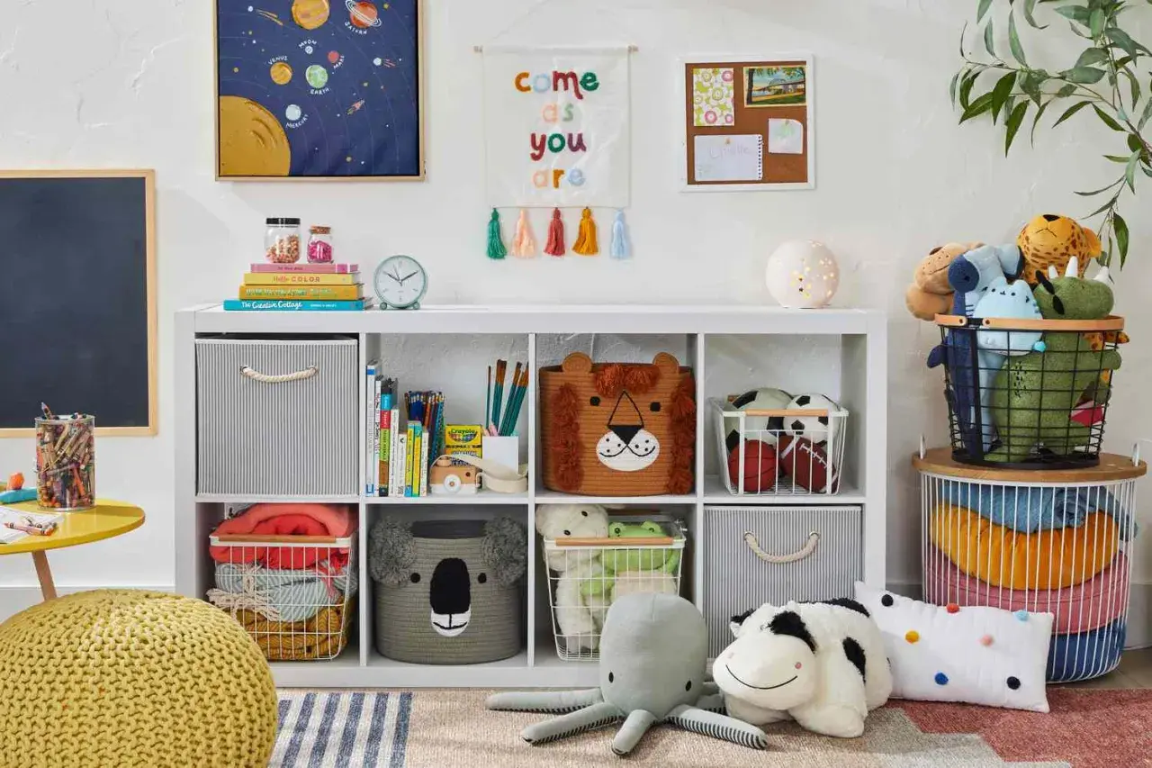 Idée petite chambre bébé : étagère blanche remplie de jouets, paniers animaux, coussins, et décorations murales colorées.