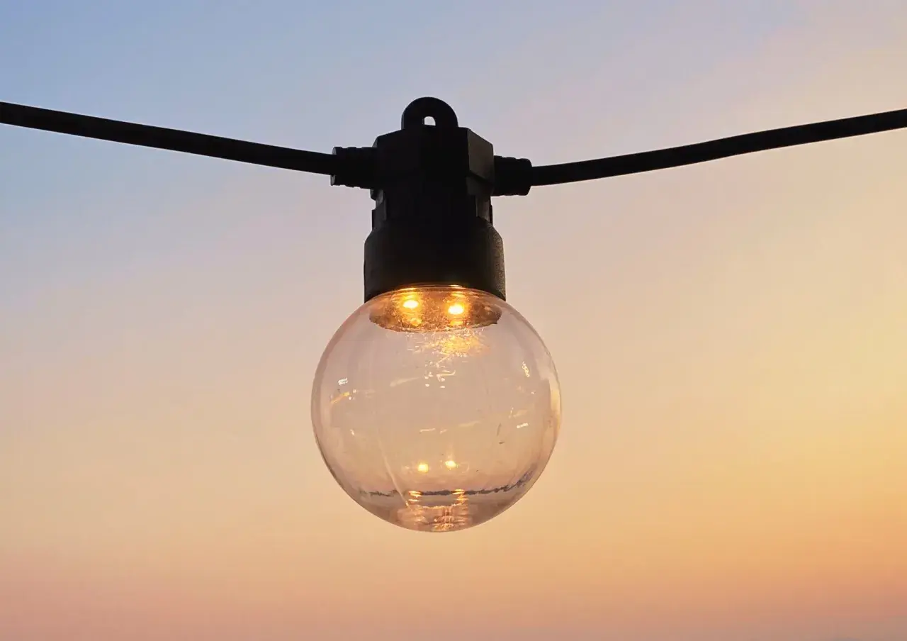 Une ampoule ronde, éclairée, suspendue à un fil noir, se reflète dans le ciel doux du soir. IP 45.