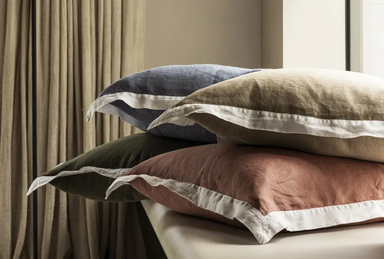 Pile de coussins aux couleurs douces, en matière drap de lin, avec des bordures blanches.