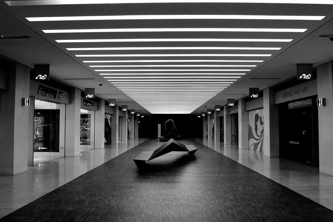 Un couloir de centre commercial, éclairci par des néons, avec une sculpture moderne au centre. Les boutiques sur les côtés sont sombres, créant un contraste saisissant.