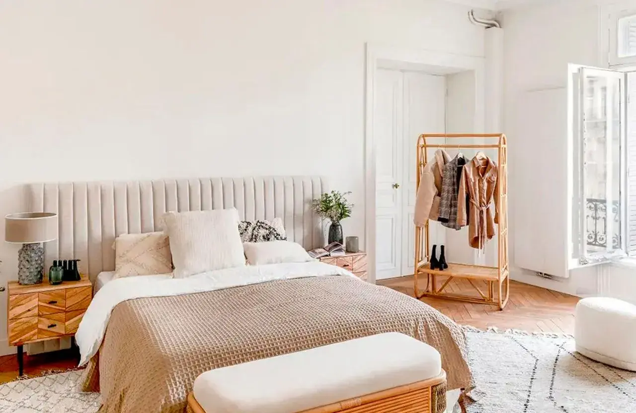 Chambre bohème scandinave avec lit douillet, table de chevet en bois, portant à vêtements en rotin et fenêtre lumineuse.