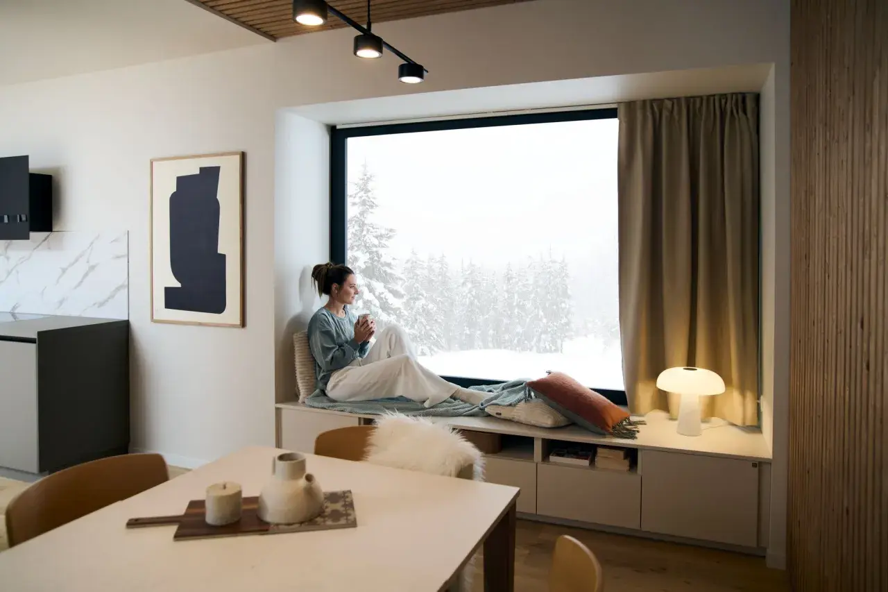Un coin lecture scandinave parfait pour admirer la neige. Une femme savoure une boisson chaude, enveloppée dans un plaid douillet.
