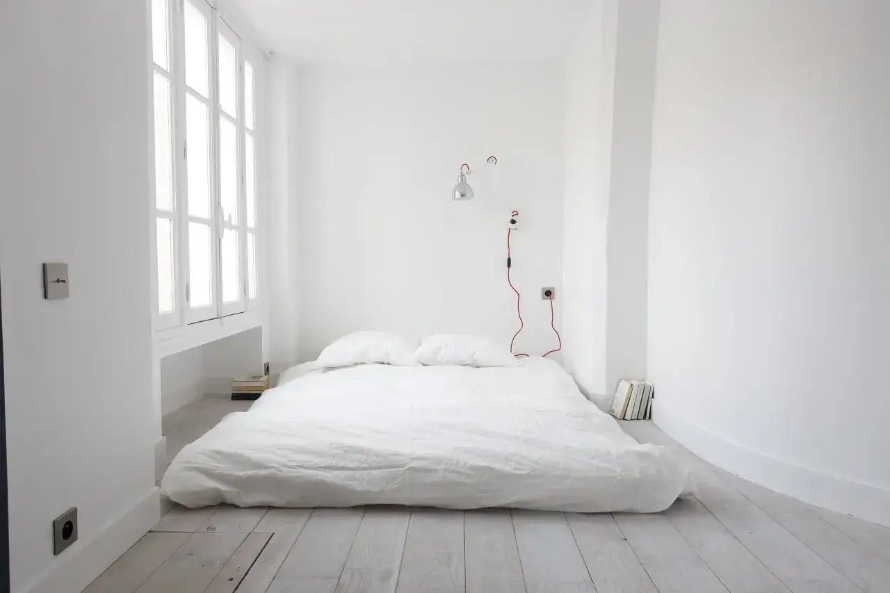 Chambre minimaliste avec un matelas au sol sans sommier, deux oreillers, une lampe murale rouge et des livres empilés.