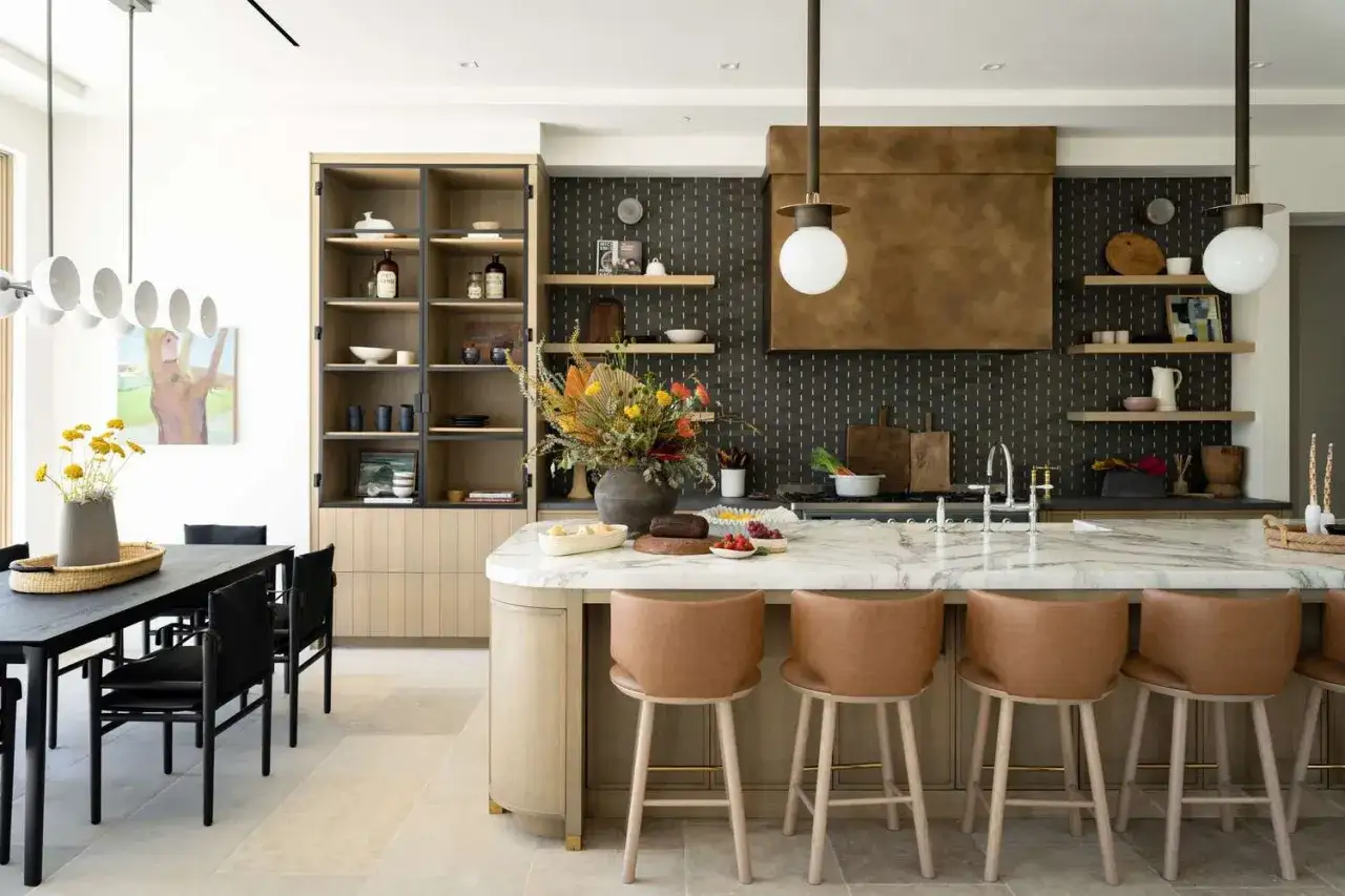 Une cuisine lumineuse couleur bois et marbre, avec un &icirc;lot central, des tabourets en cuir, une hotte dor&eacute;e et des &eacute;tag&egrave;res remplies d'objets d&eacute;coratifs.