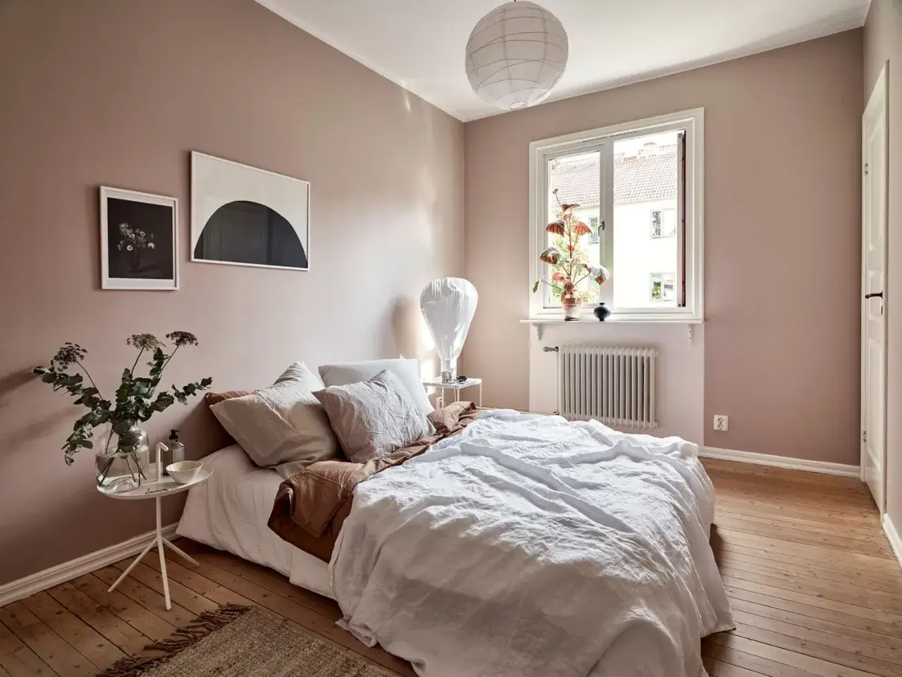 Chambre rose poudr&eacute; et bois, lit blanc douillet, table de chevet avec vase de fleurs, et deux tableaux au mur.