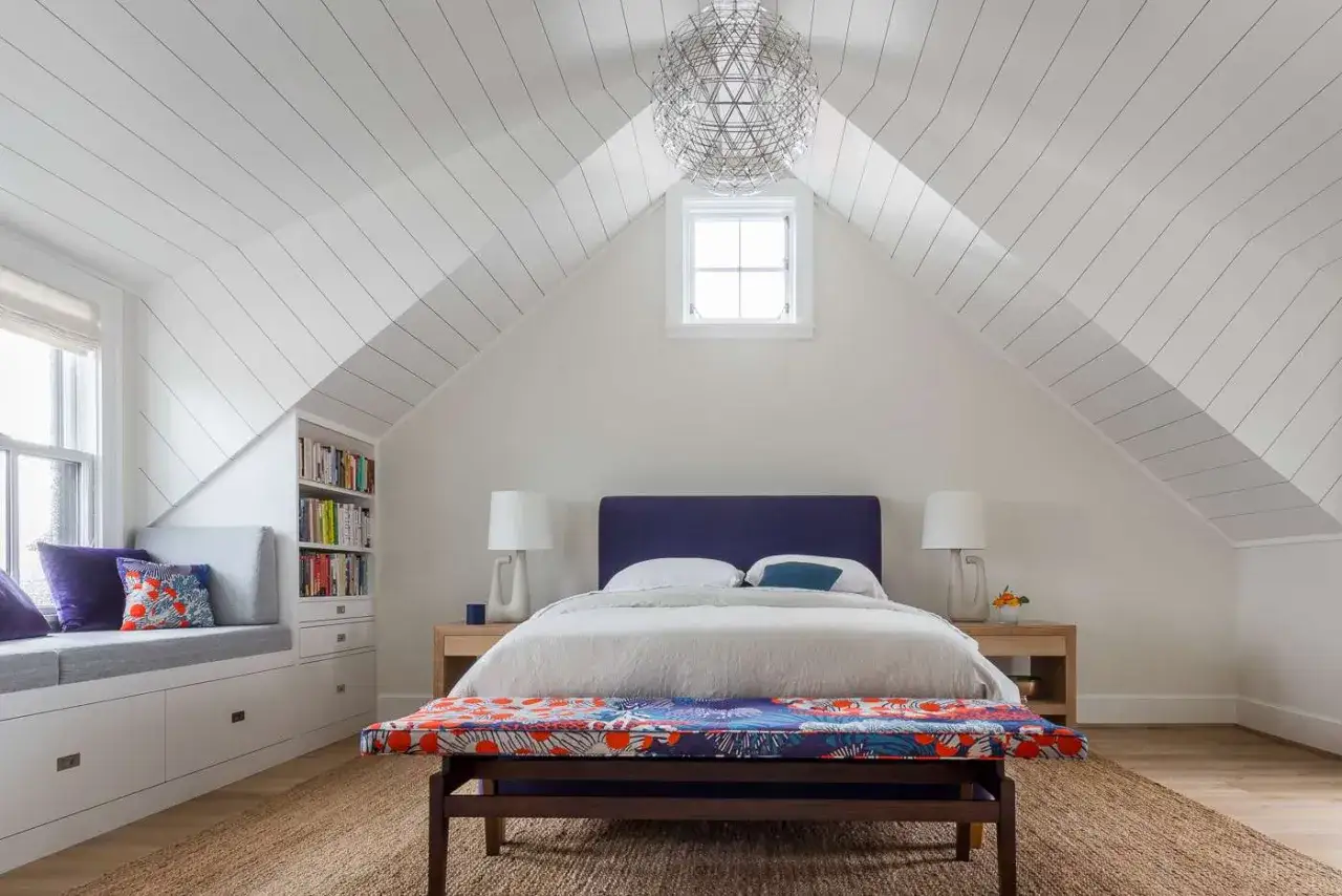 Chambre mansard&eacute;e aux murs blancs et au plafond &agrave; deux couleurs, avec un lit bleu marine et un banc color&eacute;.