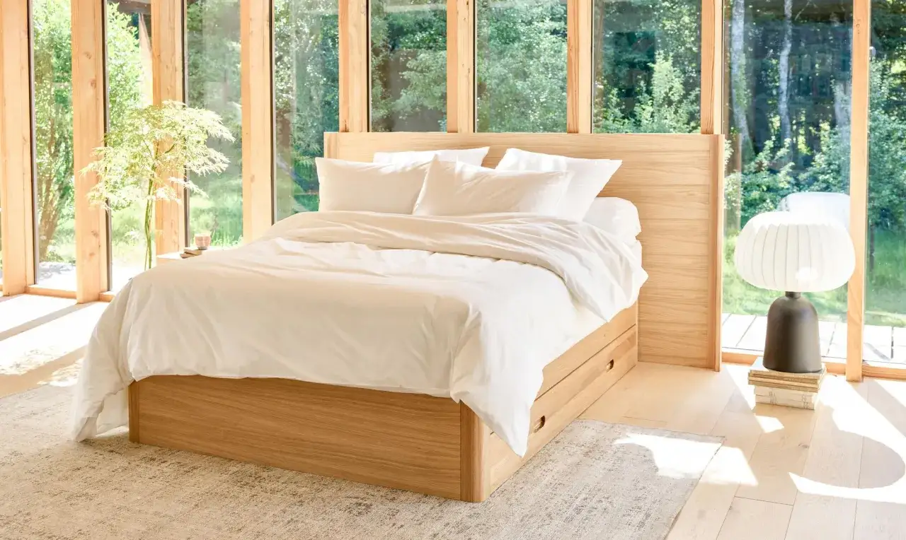 Une chambre minimaliste avec un lit en bois, des draps blancs immacul&eacute;s et de grandes fen&ecirc;tres donnant sur la nature.