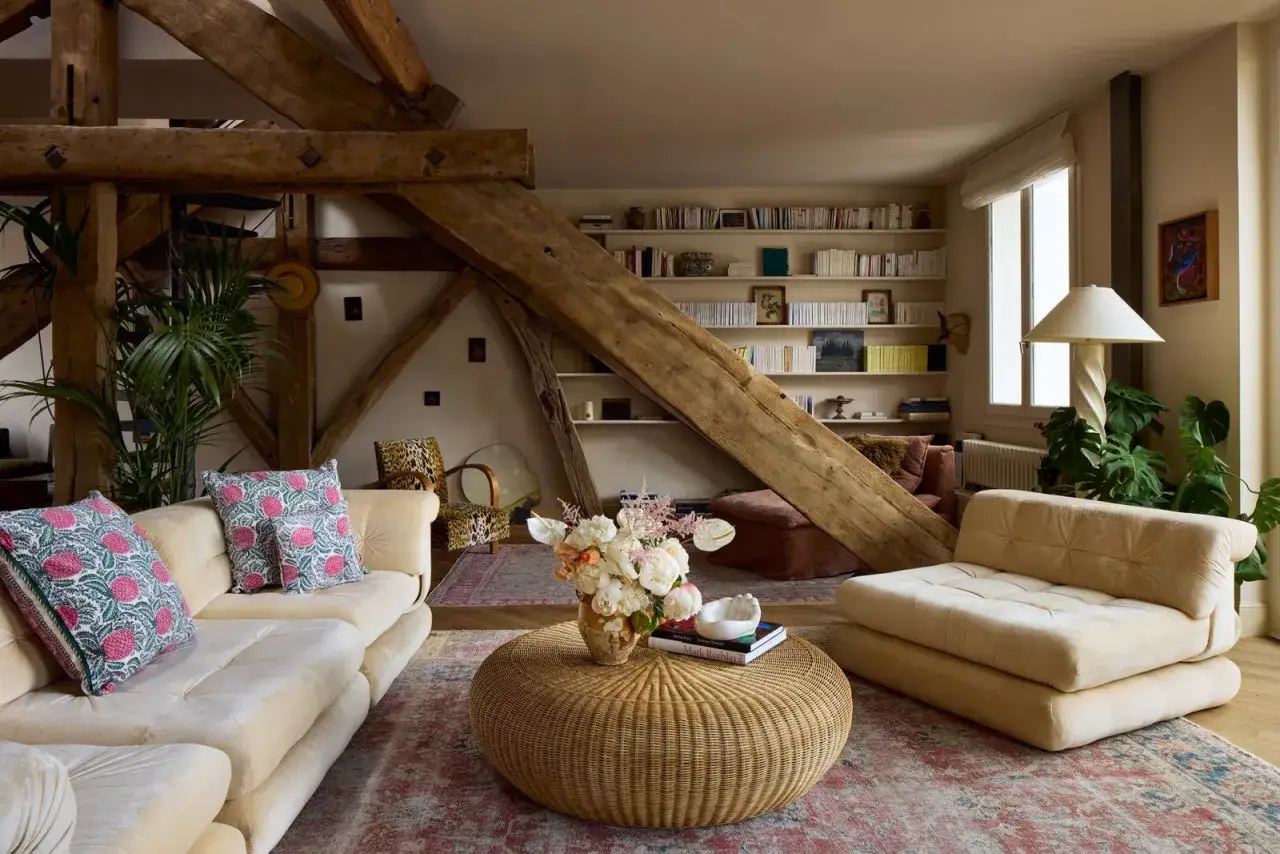 Un salon cocooning sous les poutres apparentes, avec canapé, fauteuil, table basse en rotin et bibliothèque remplie.