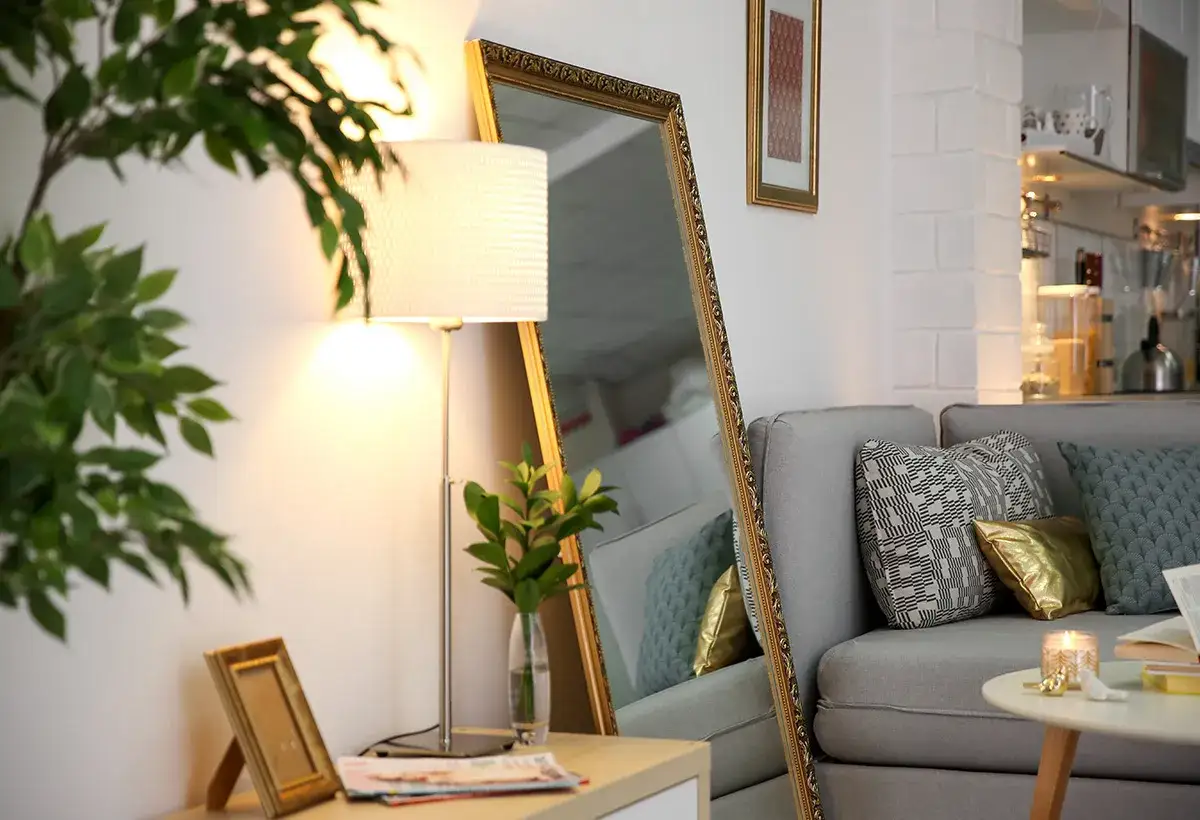 Un grand miroir derri&egrave;re un canap&eacute; gris, refl&eacute;tant une lampe et des coussins d&eacute;coratifs. Une plante verte ajoute une touche de fra&icirc;cheur.