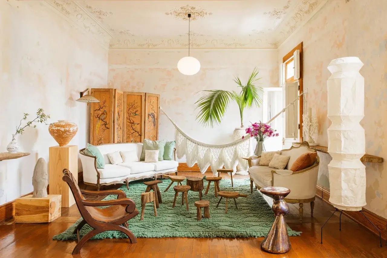 Salon avec une d&eacute;co boh&egrave;me chic : canap&eacute;s blancs, hamac en macram&eacute;, tapis vert, meubles en bois brut et paravents orn&eacute;s.