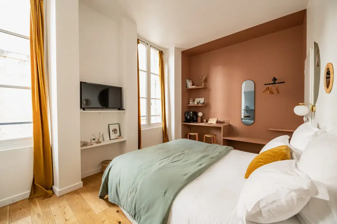 Chambre d'un petit appartement moderne : lit douillet, mur couleur terre cuite, étagères minimalistes, TV et rideaux ocre.