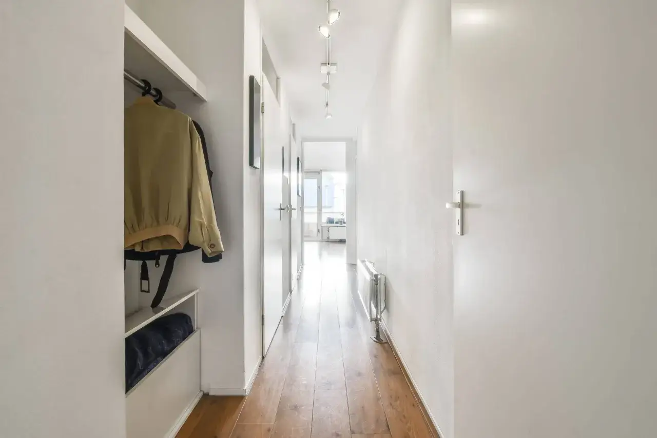 Un long et étroit couloir baigné de lumière naturelle. Un vestiaire avec une veste jaune est visible à gauche.