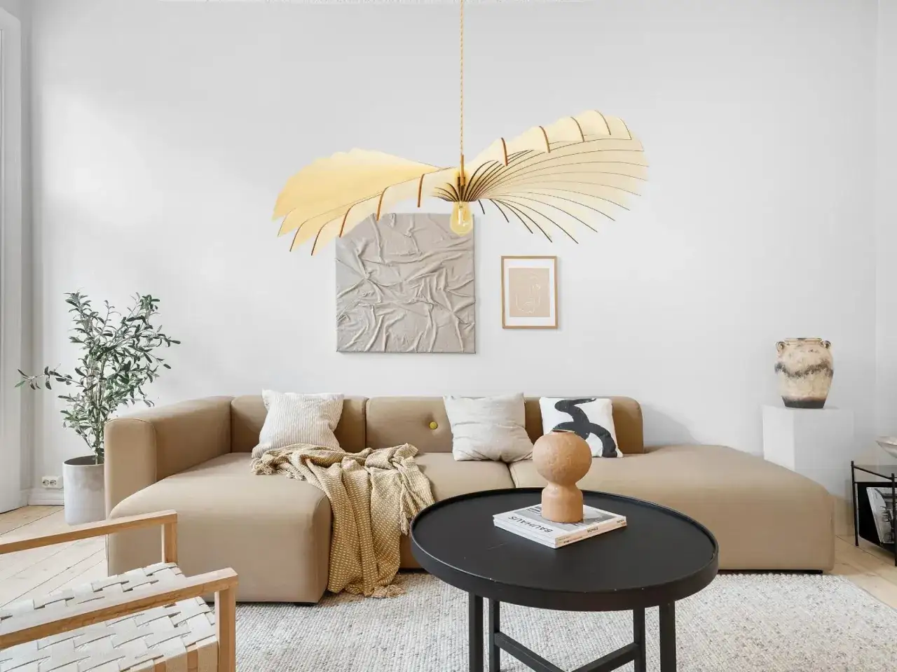 Un salon moderne avec un canap&eacute; beige, une table basse noire et une plante verte. La tendance luminaire est marqu&eacute;e par un lustre en bois sculptural au-dessus du canap&eacute;.