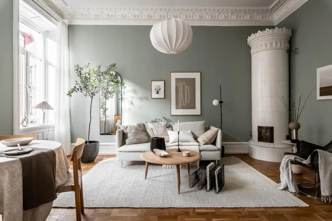 Salon avec murs vert sauge et sol en parquet. Un canapé gris clair est placé sur un tapis blanc. Quelle couleur de mur avec un sol gris foncé ? Ce salon est une inspiration.