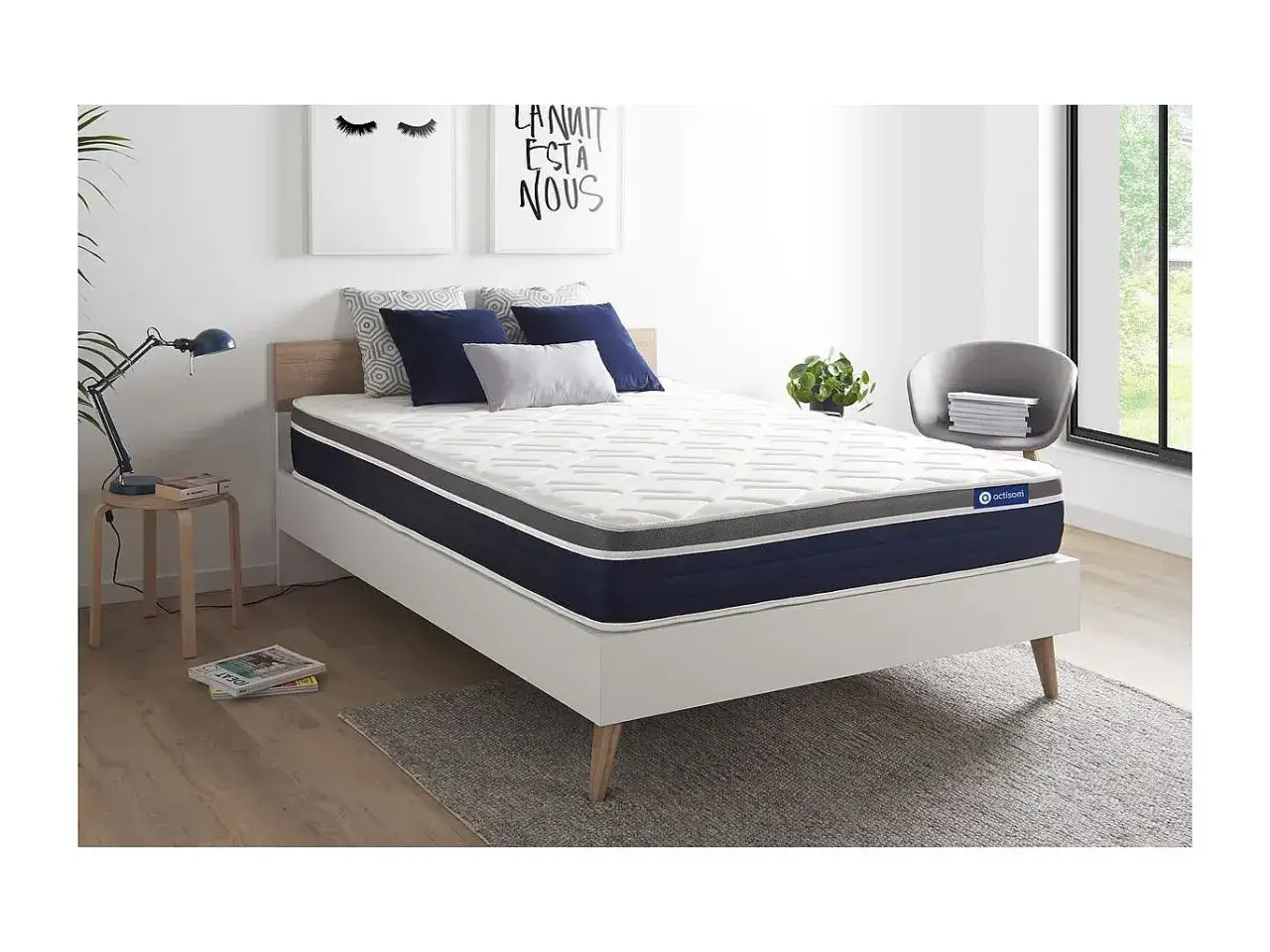 Un matelas confortable, peut-&ecirc;tre en latex ou m&eacute;moire de forme, est pr&eacute;sent&eacute; sur un lit blanc, dans une chambre moderne avec des coussins bleus et gris.