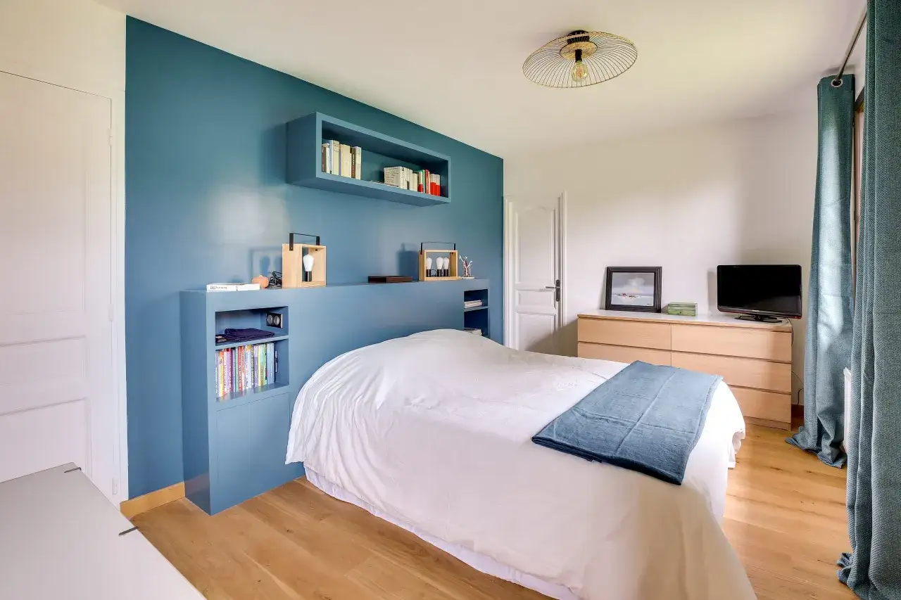 Chambre bleu et beige avec un lit blanc, des étagères bleues remplies de livres et une commode en bois clair.