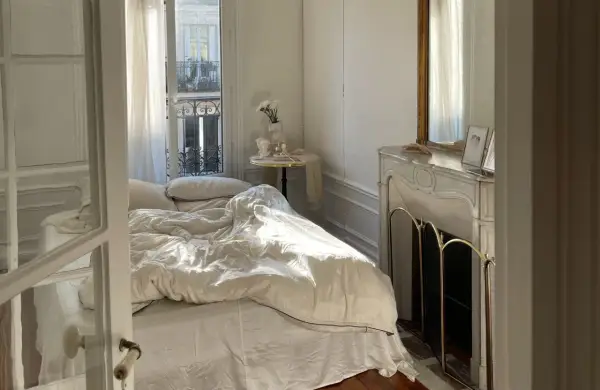 Une petite chambre cocooning baignée de soleil. Le lit défait invite au repos, une cheminée ornée de bibelots et un miroir doré complètent ce tableau paisible.
